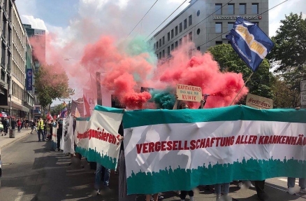 Demo-Block des Bündnisses "Profite Schaden Ihrer Gesundheit" auf der Demo zum TVE-Streik in NRW. Auf dem Frontbanner steht "Vergesellschaftung aller Krankenhäuser", außerdem werden Rauchtöpfe gezündet.